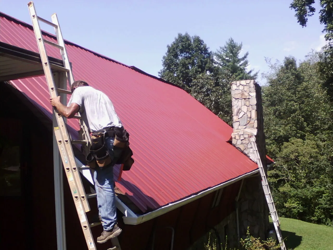 Roofer on a ladder beside a red metal roof and stone chimney — Soffit & Fascia Repair by Roofing Brothers in Jasper